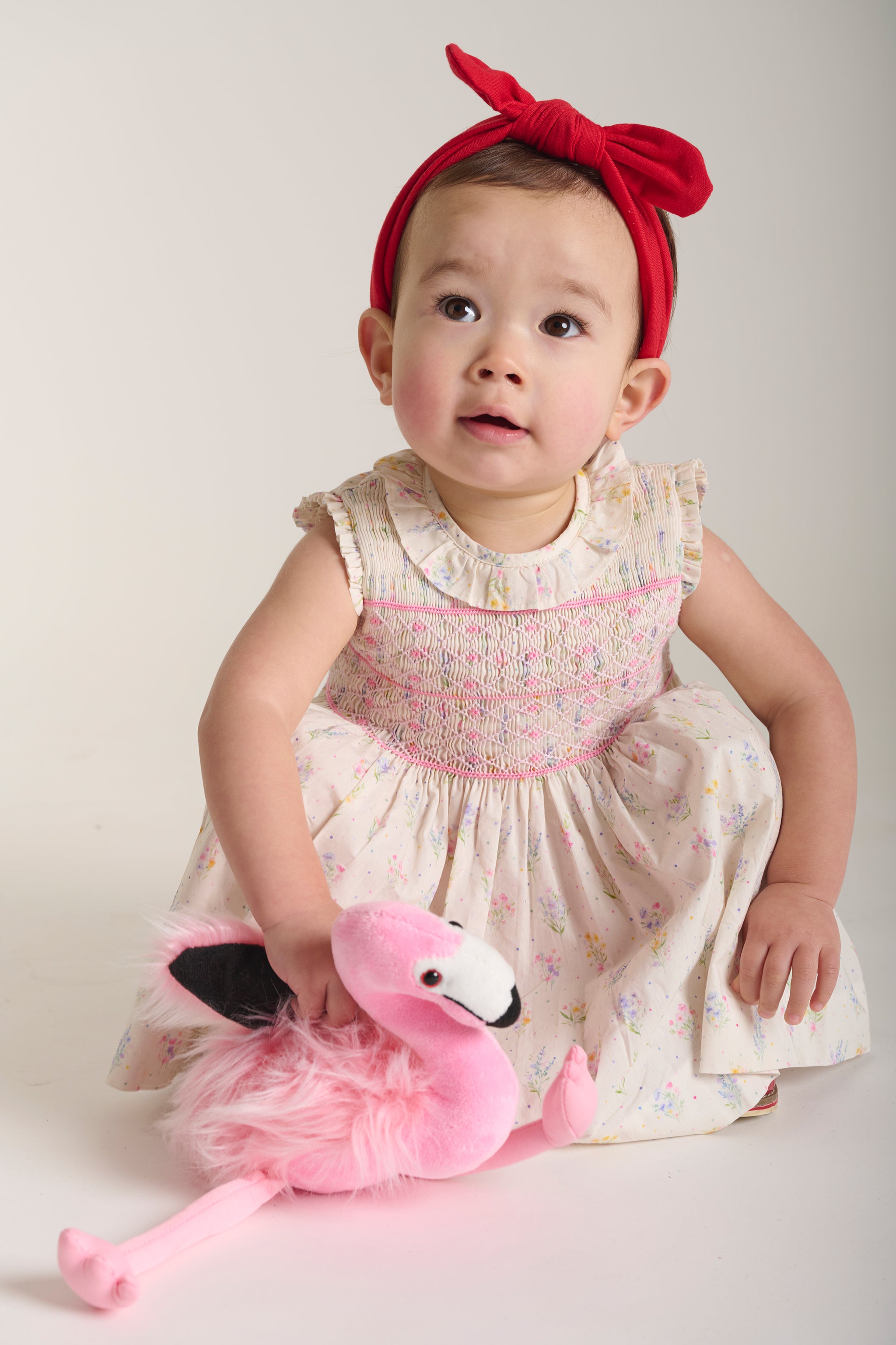 Baby in a floral dress with a red headband holding a pink plush flamingo toy.