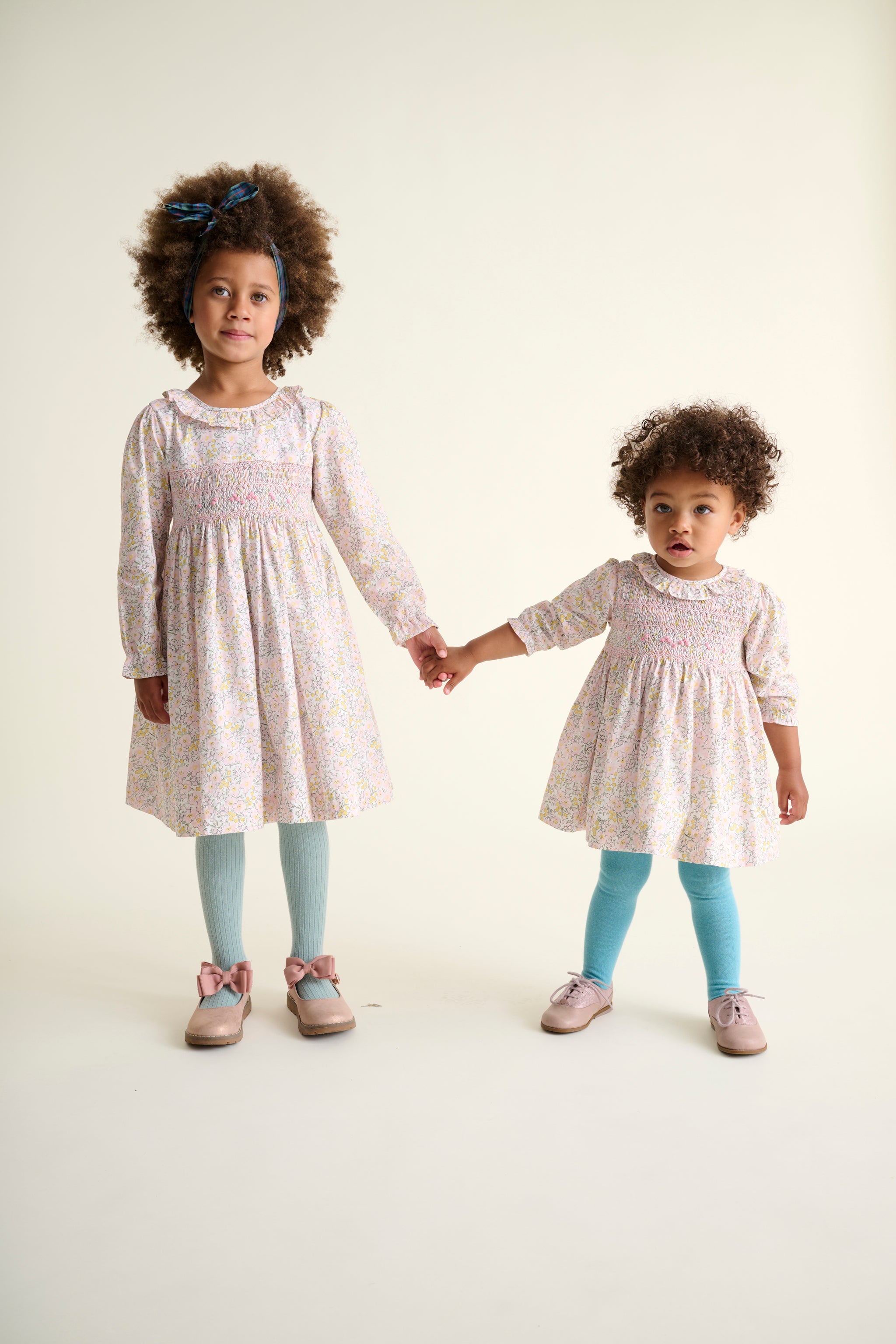 Two young girls holding hands, wearing matching dresses and light blue tights on a plain background