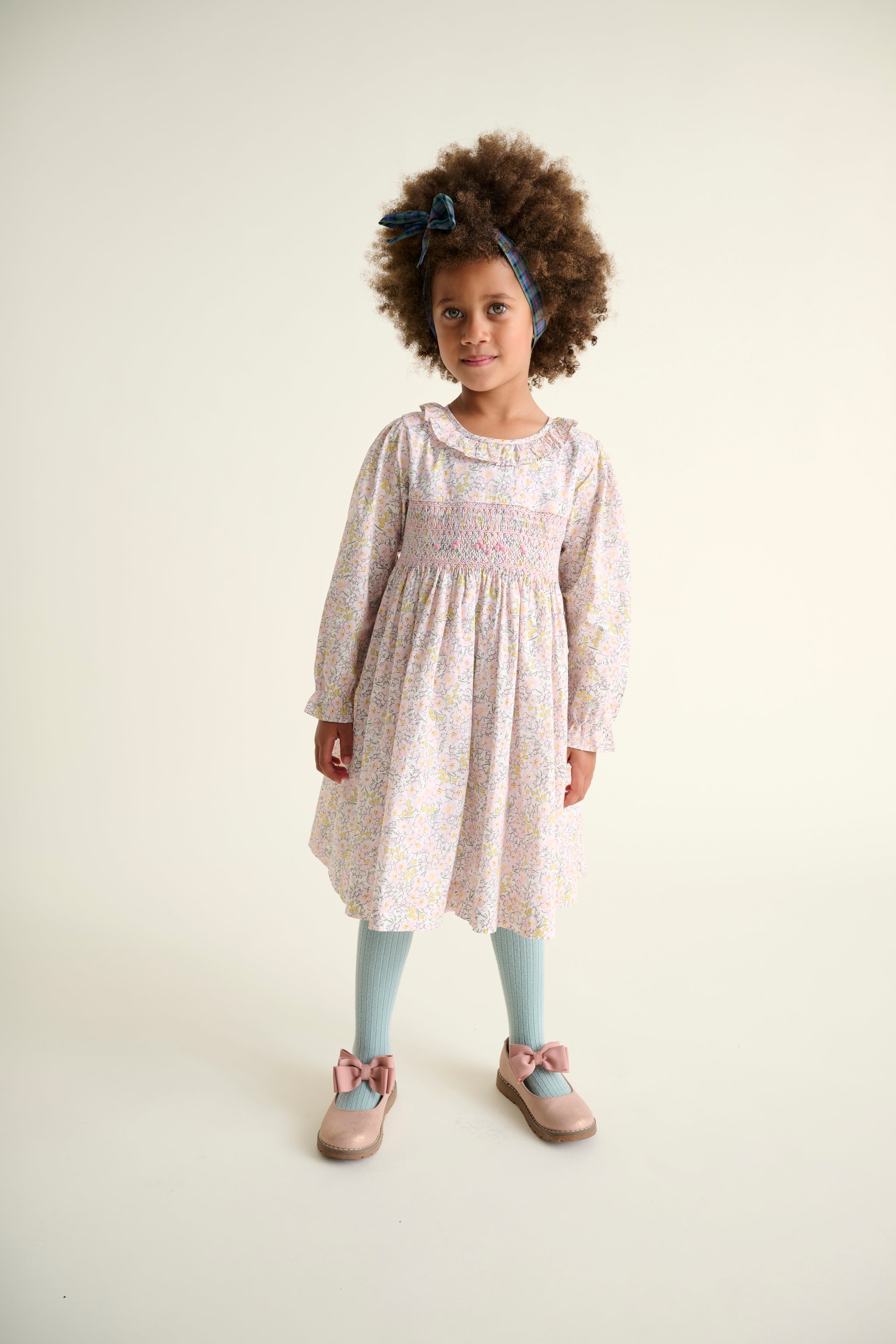 Young girl wearing a patterned smocked dress with light blue tights and shoes on a white background