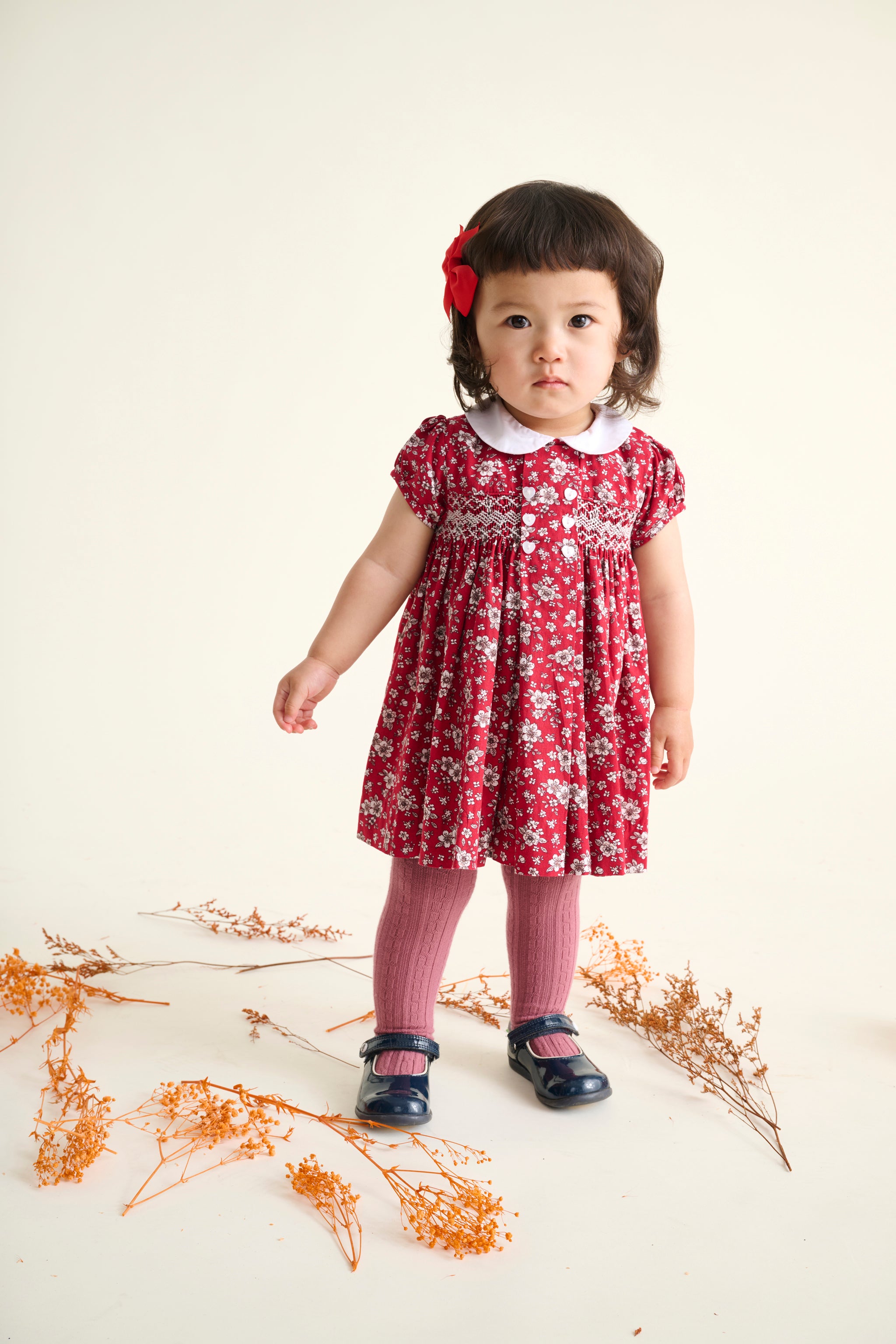 toddler girl in a festive red floral dress standing on a white background with dried flowers.