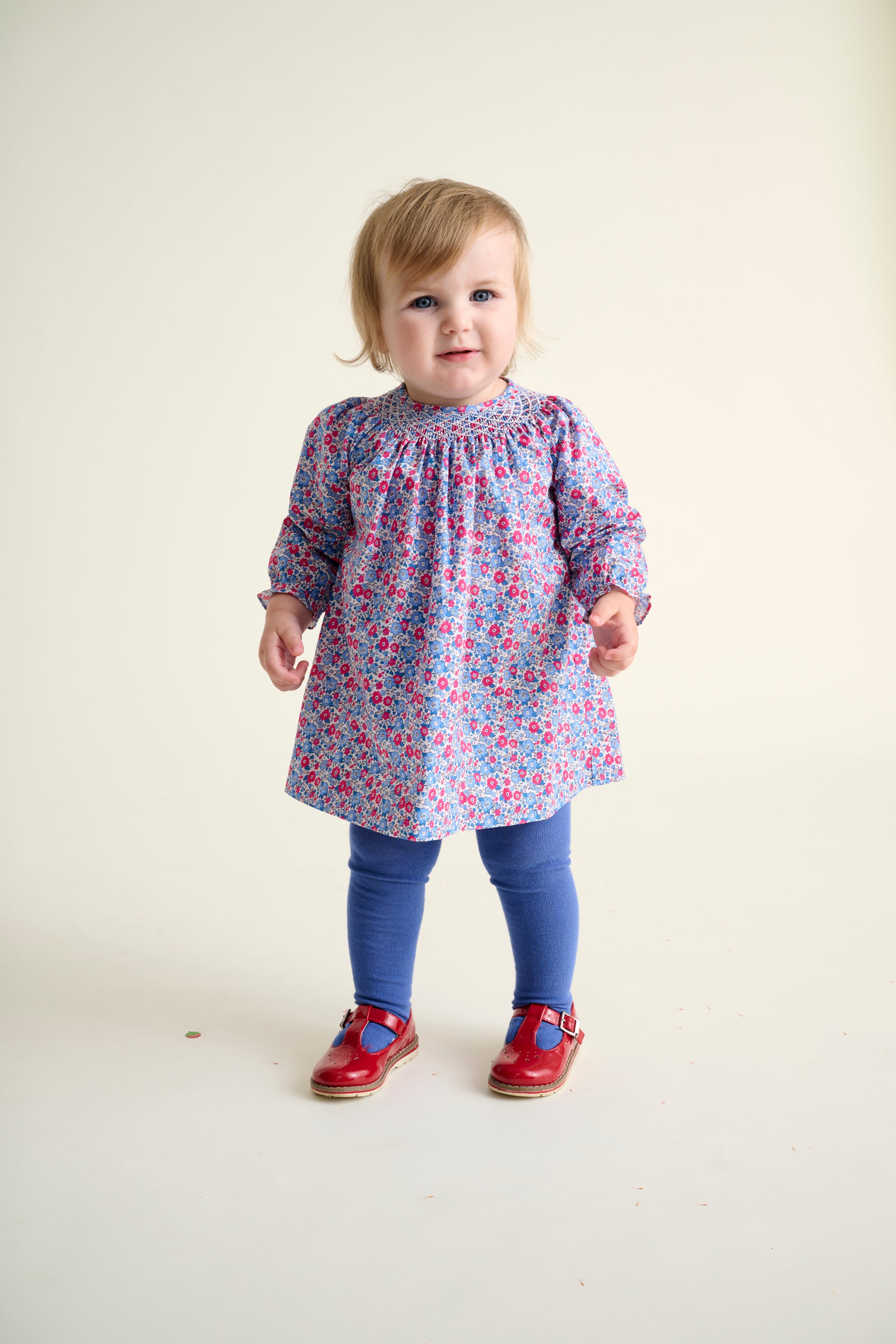 toddler girls wearing a floral smock dress and blue tights with red shoes on a white background