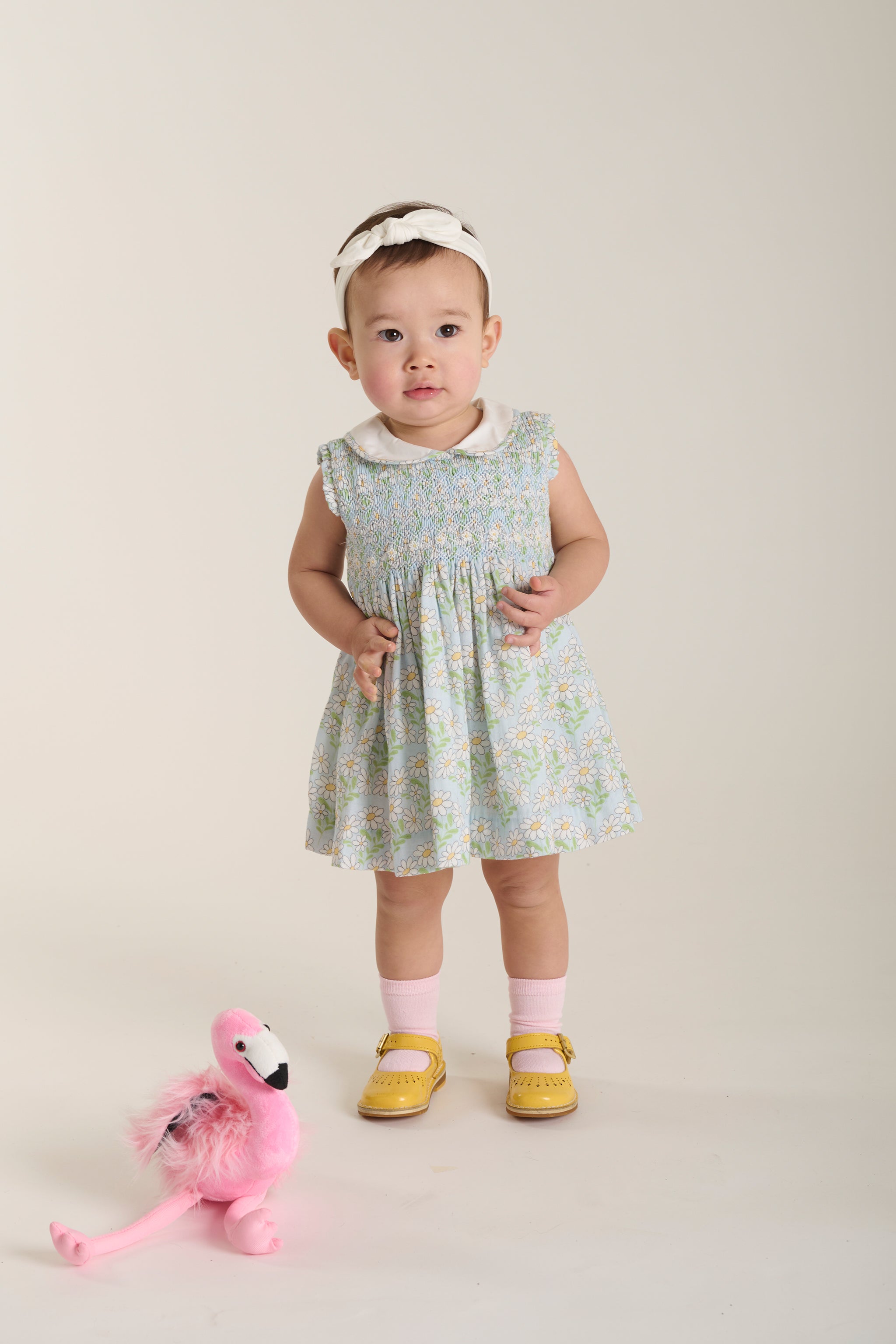 Baby in a floral dress with a pink flamingo toy on a beige background
