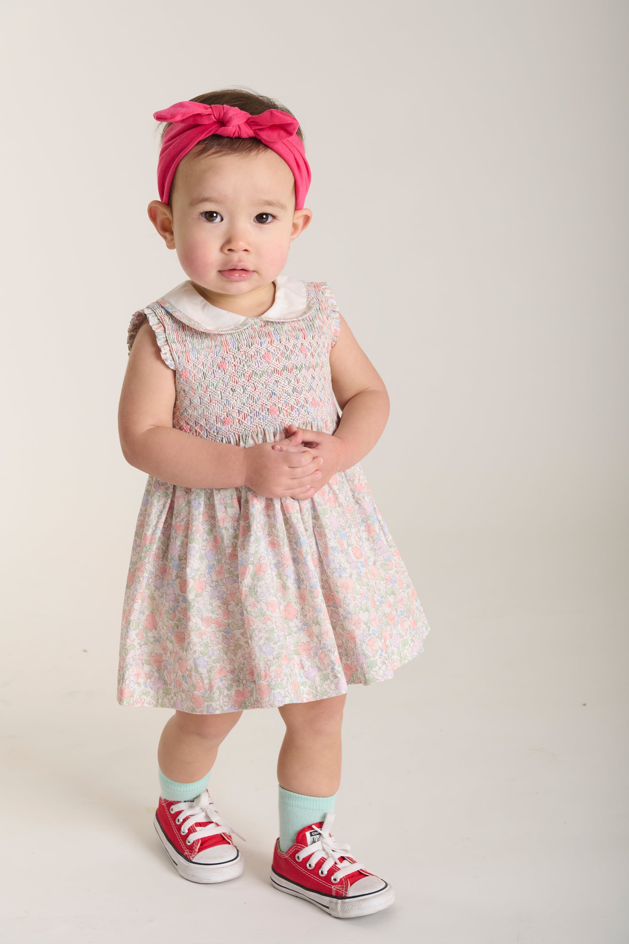 Baby wearing a floral smock dress with red shoes and a pink headband on a white background