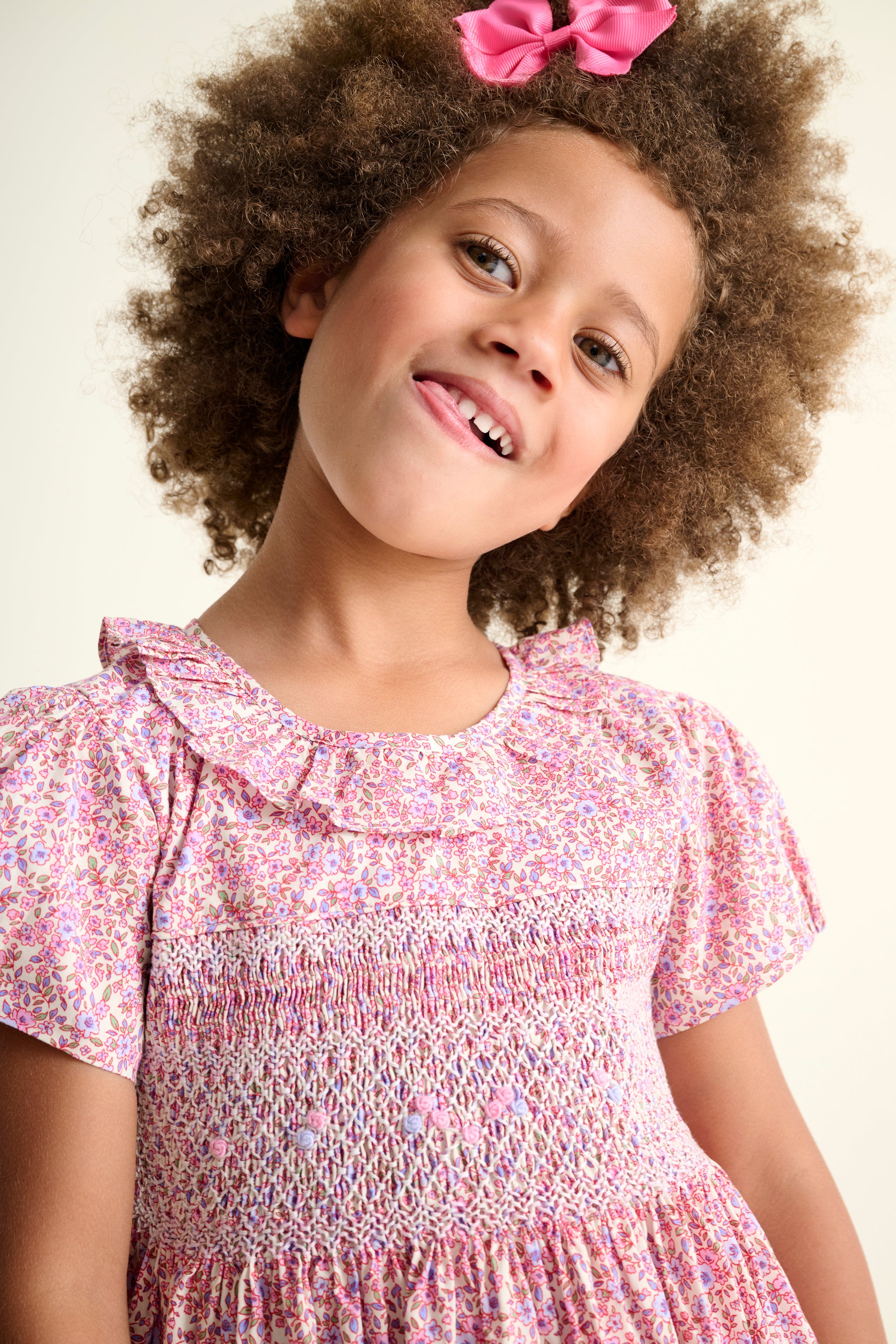 Young girl wearing a pink floral hand smocked dress with a bow in her hair against a plain background