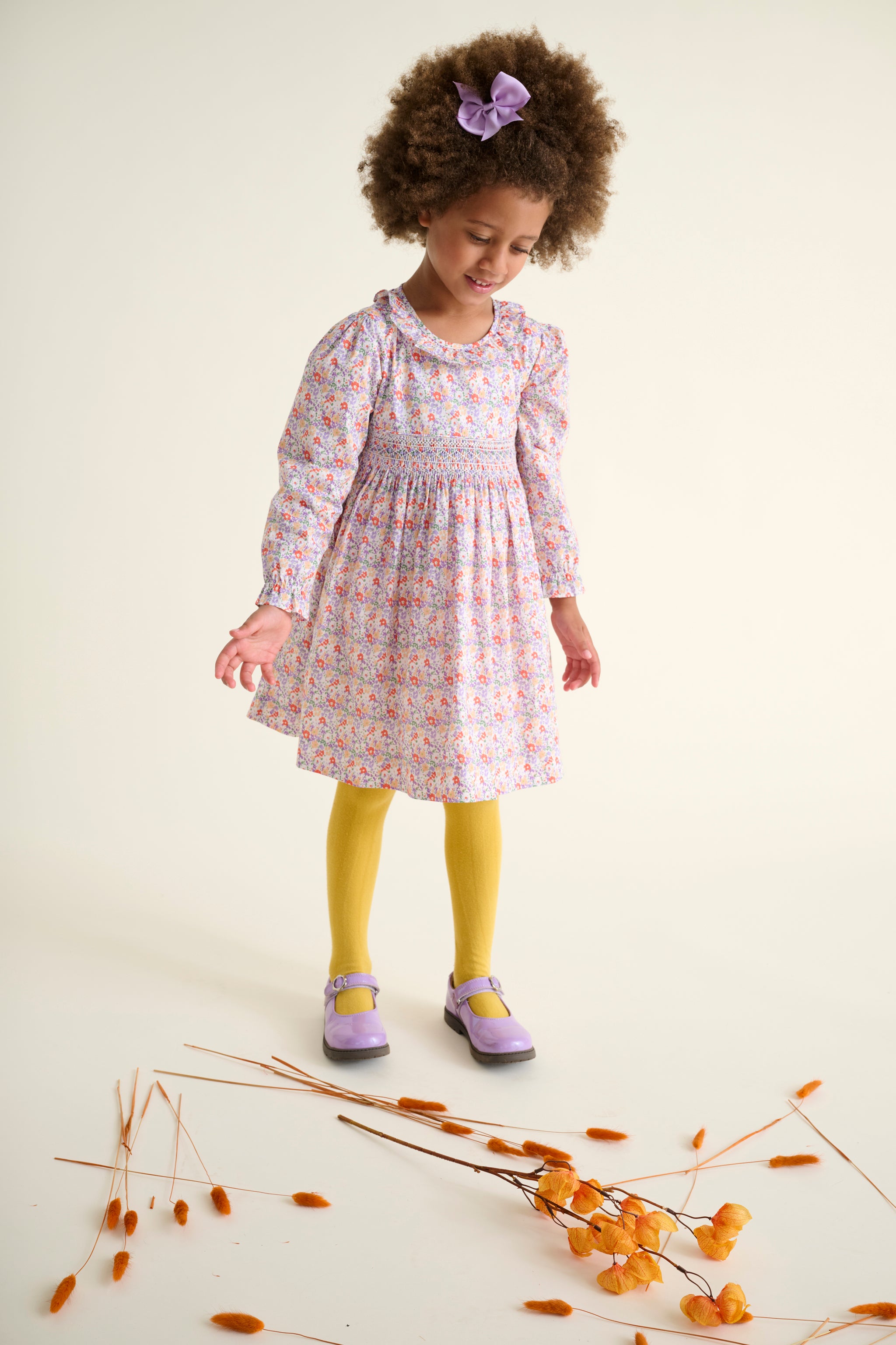 Young girl in a floral dress with yellow tights and purple shoes standing on a white background with dried flowers.