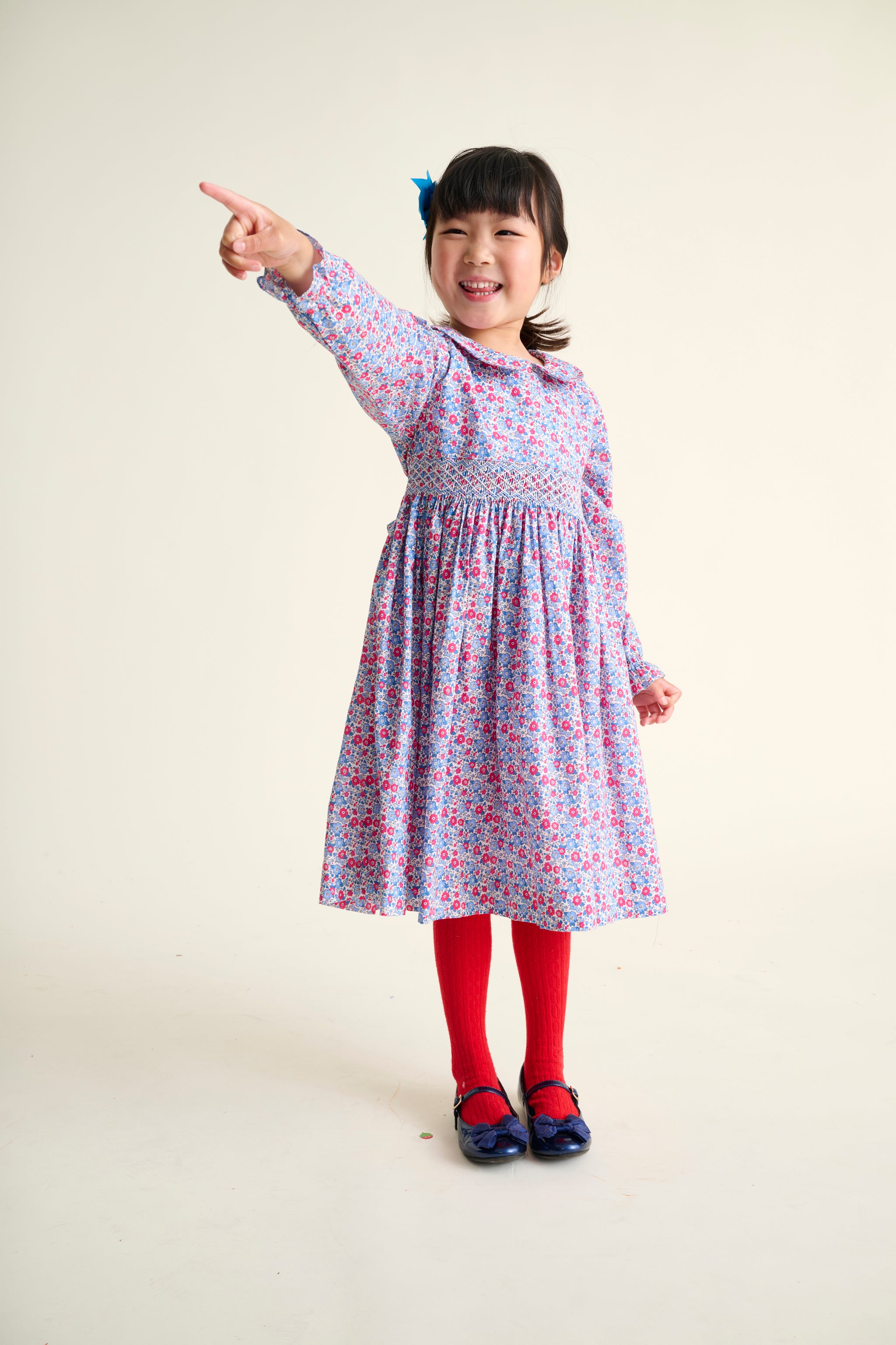 Young girl wearing a floral dress pointing towards the camera on a plain background