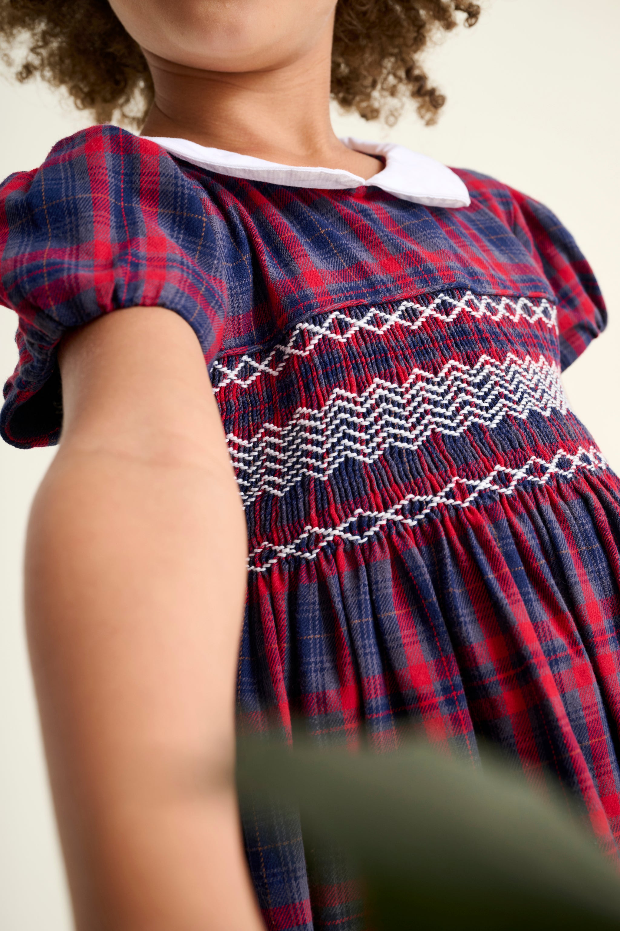 Child wearing a red and blue plaid dress with white smocking details on a plain background