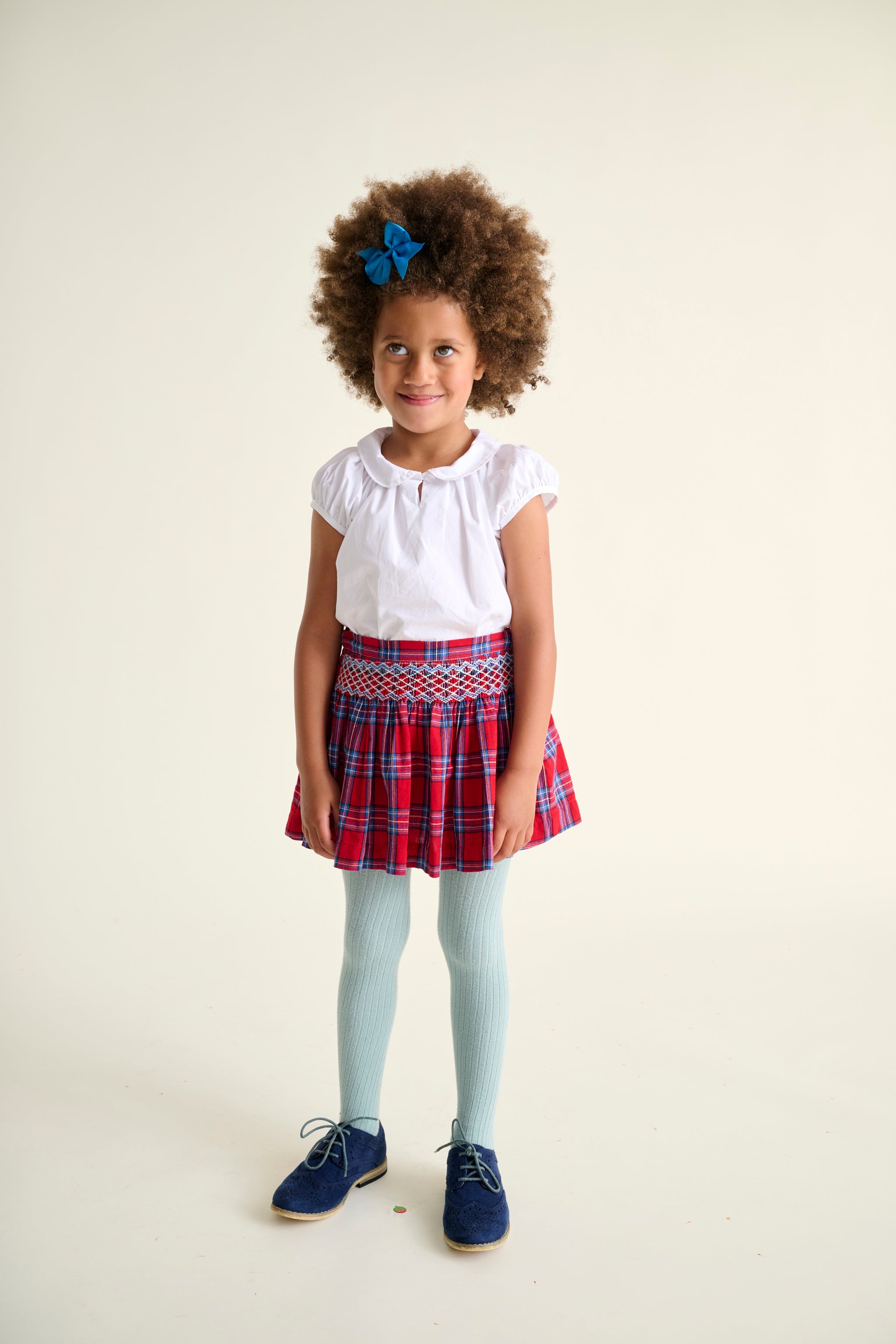 Young girl wearing a white top, red plaid skirt, and blue shoes on a plain background