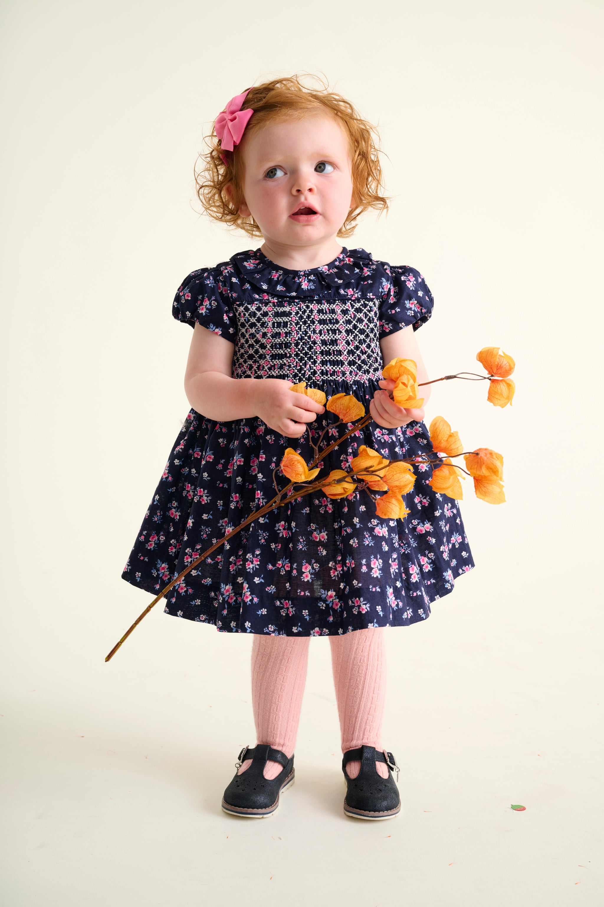 Toddler girl in a floral dress holding orange flowers against a white background