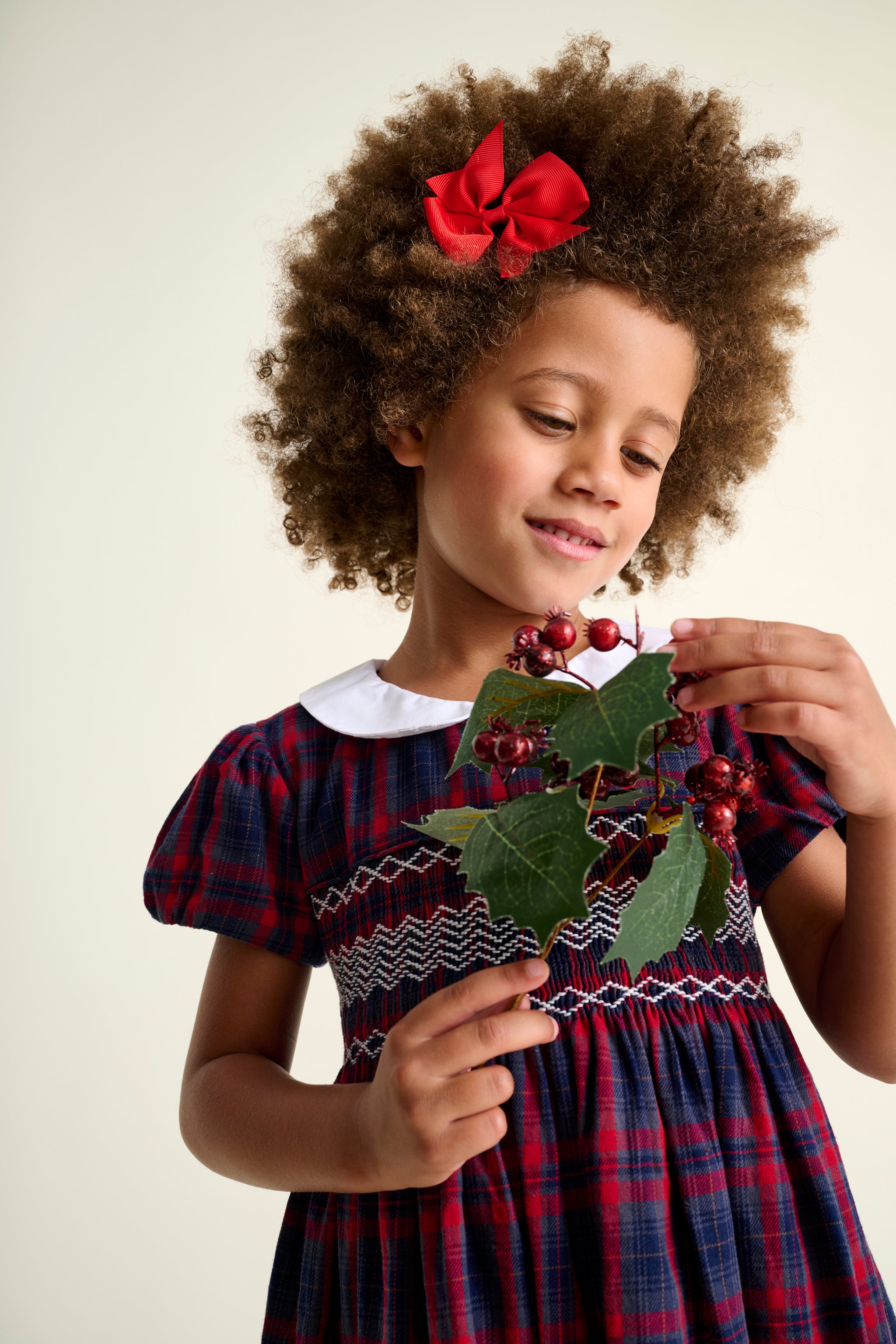 Young girl in a plaid dress holding a decorative wreath with red berries and green leaves.