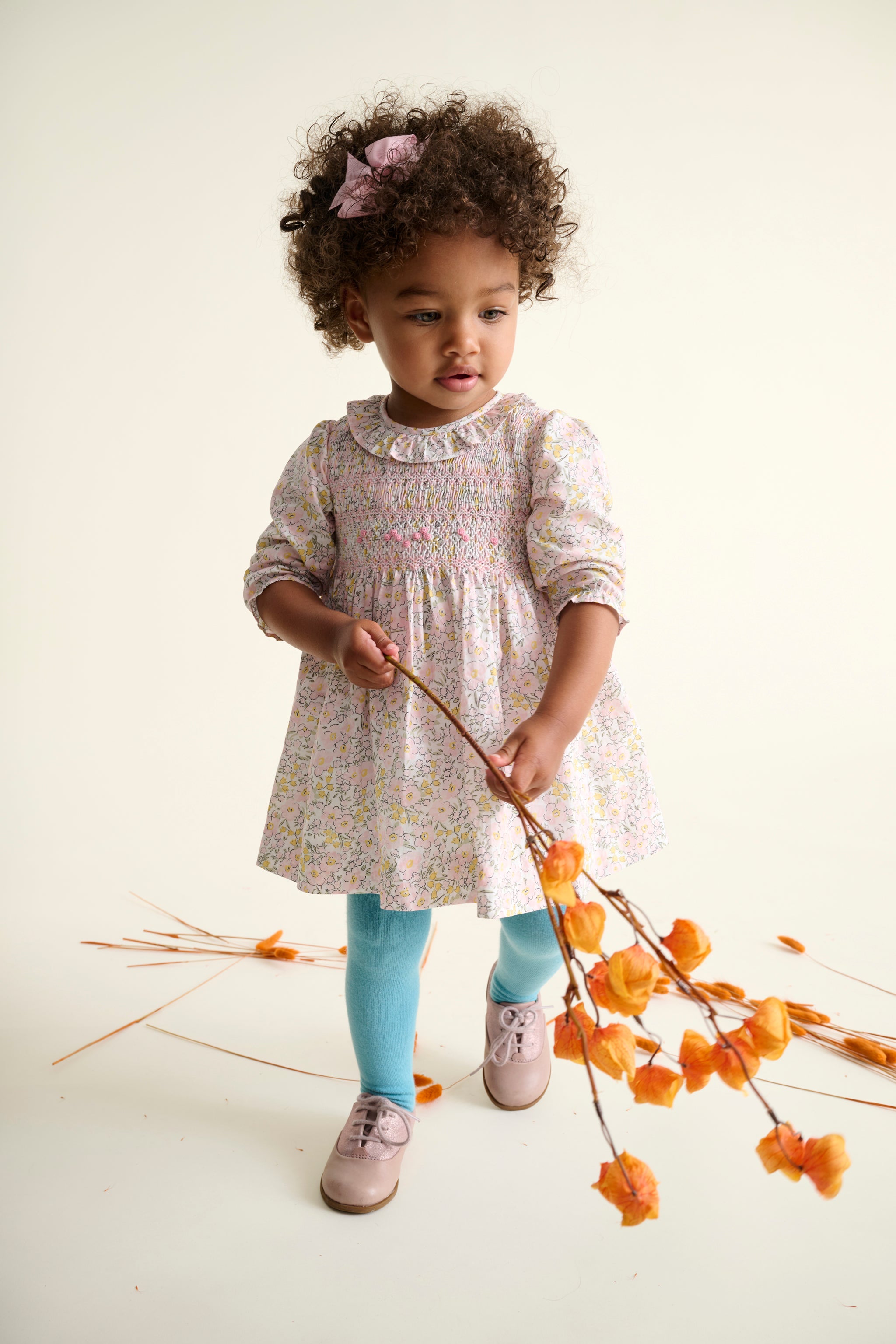 toddler girl in a floral dress holding a branch with orange flowers on a white background