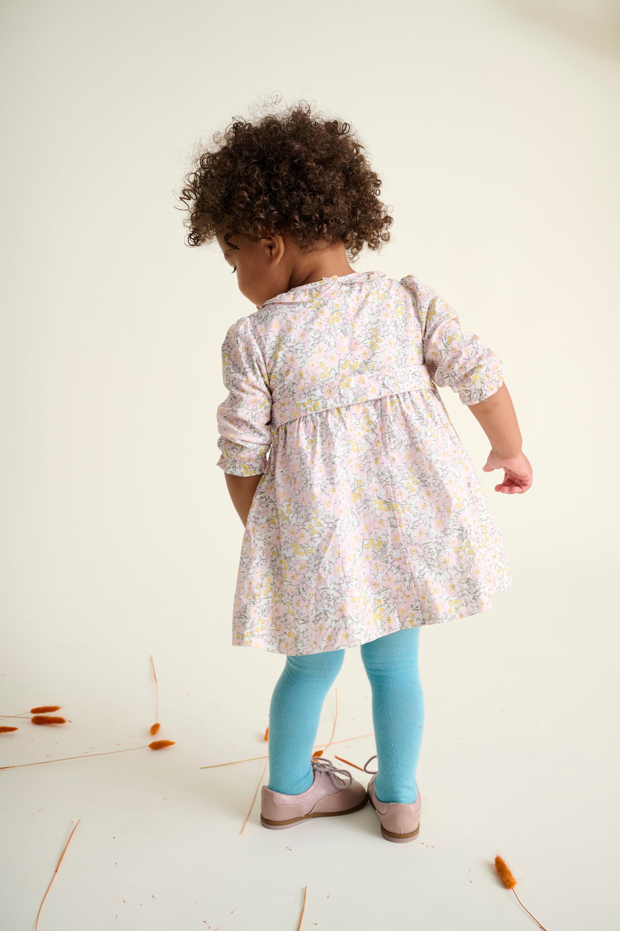 back of a child wearing a floral dress and blue leggings on a white floor.