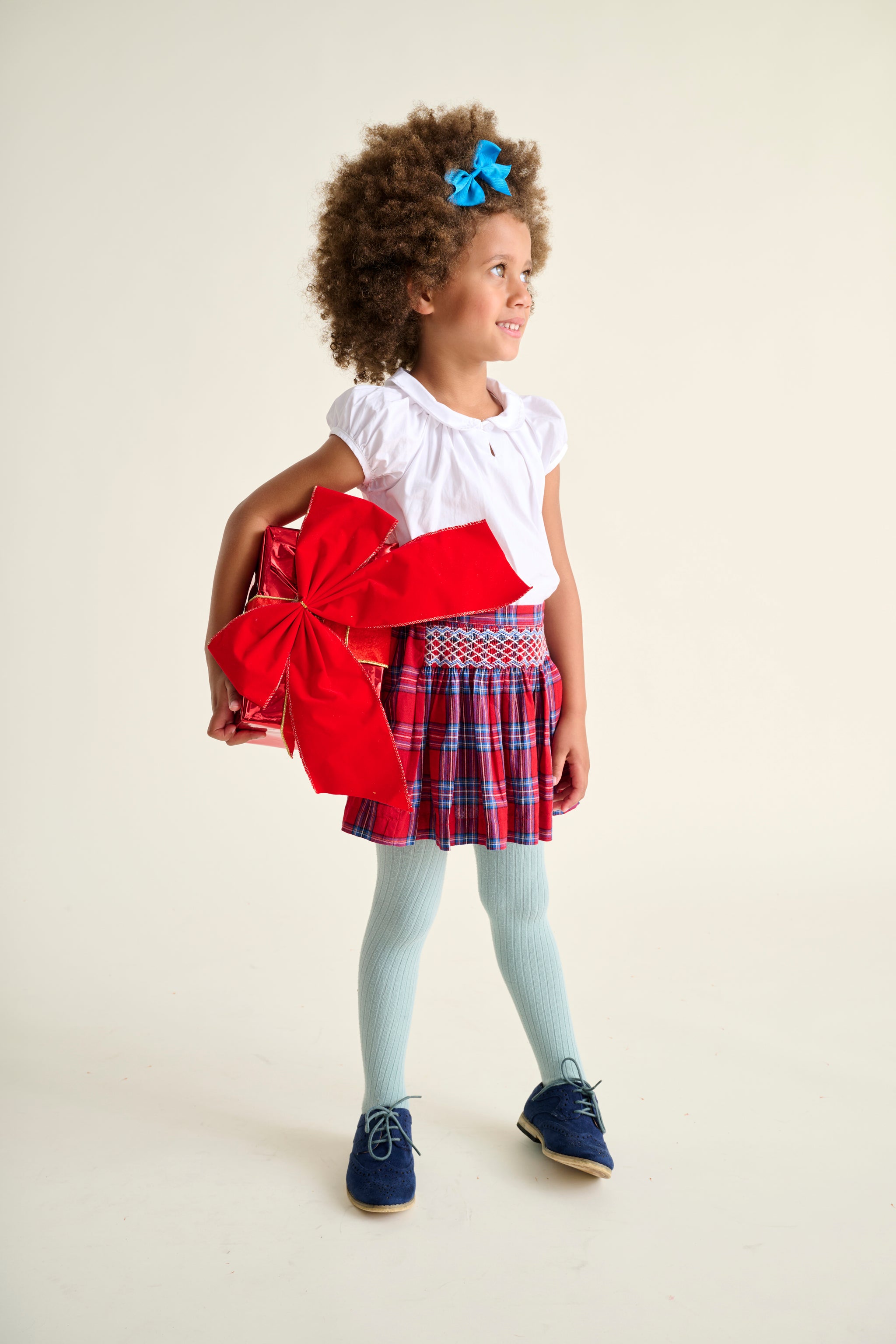 Young girl holding a red bow, wearing a white top and a hand-smocked plaid skirt holding a big gift