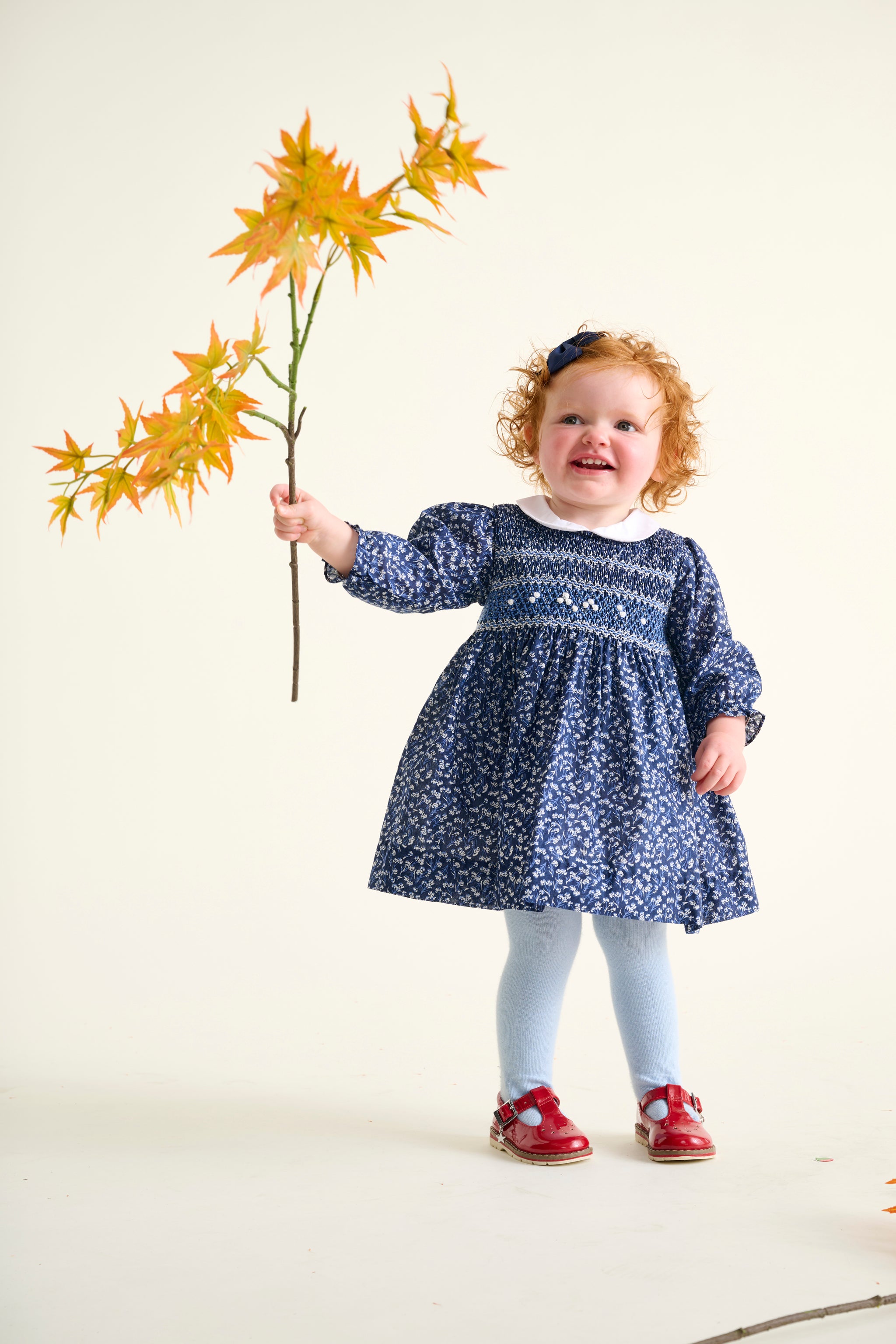 Toddler in a blue floral hand-smocked dress holding a branch with yellow leaves on a white background