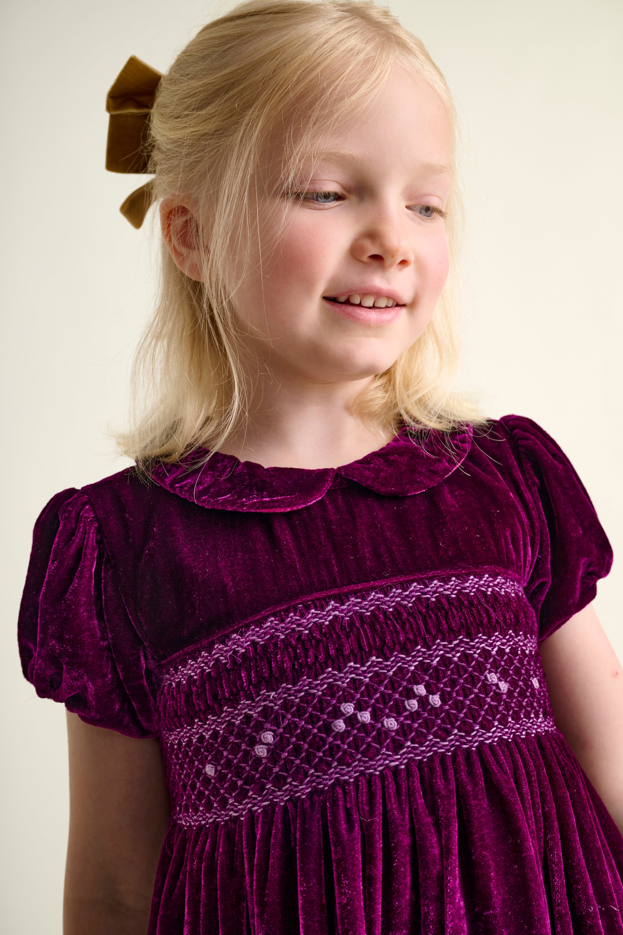 Young girl wearing a burgundy velvet dress with smocking against a plain background