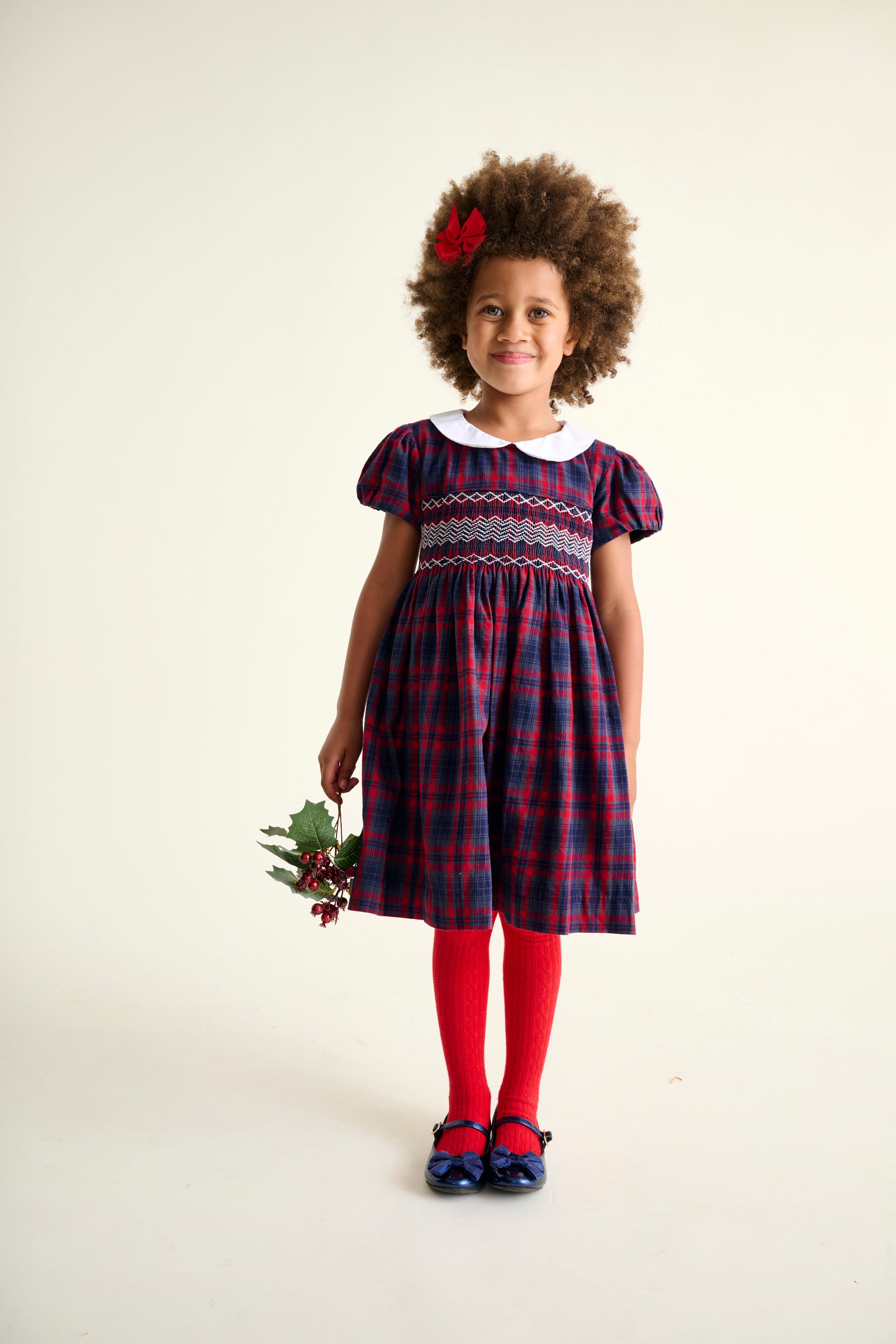 Young girl wearing a navy and red tartan hand-smocked dress with red tights and shoes, holding a  mistletoe