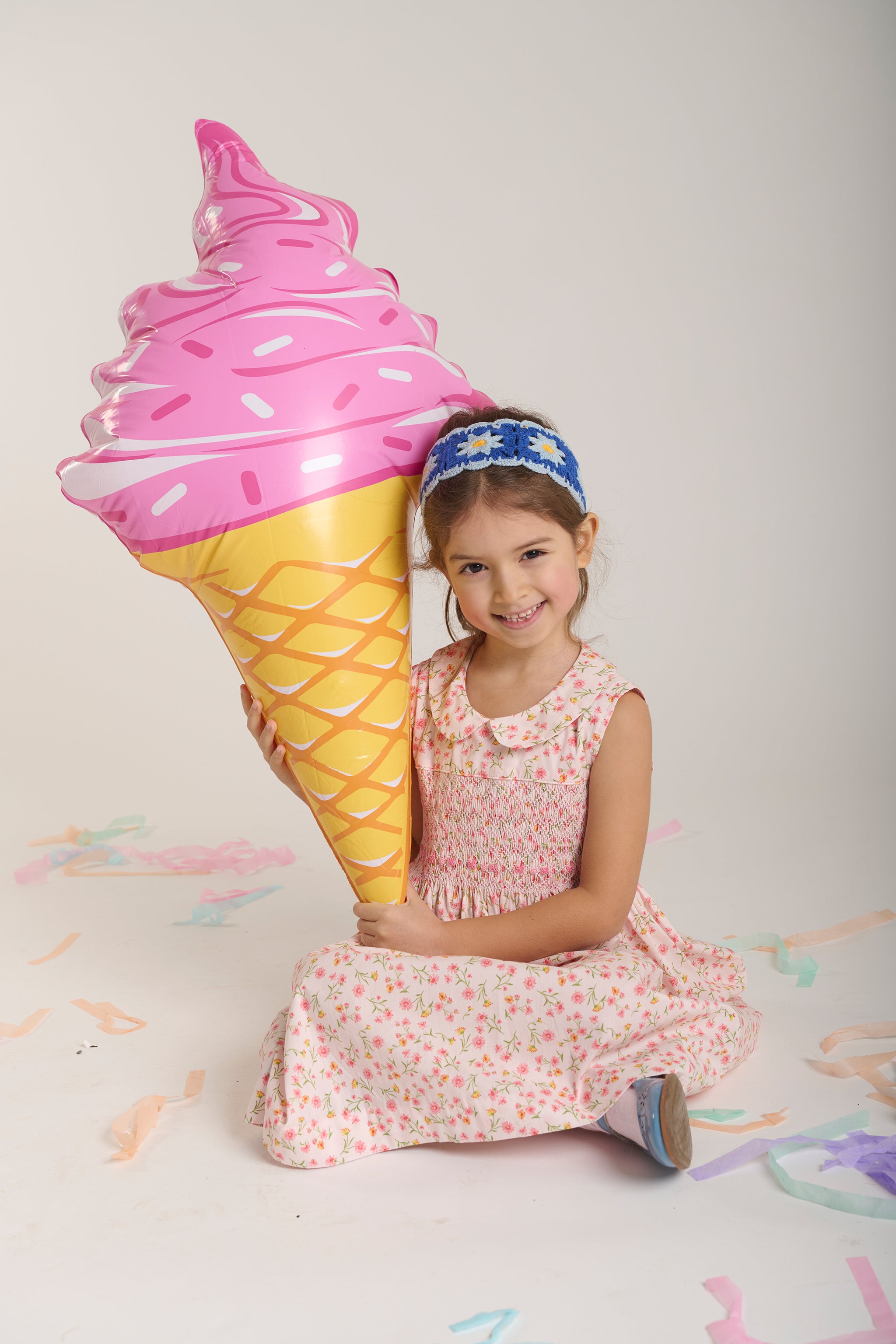 Child holding a large inflatable ice cream cone against a plain background in a smocked dress
