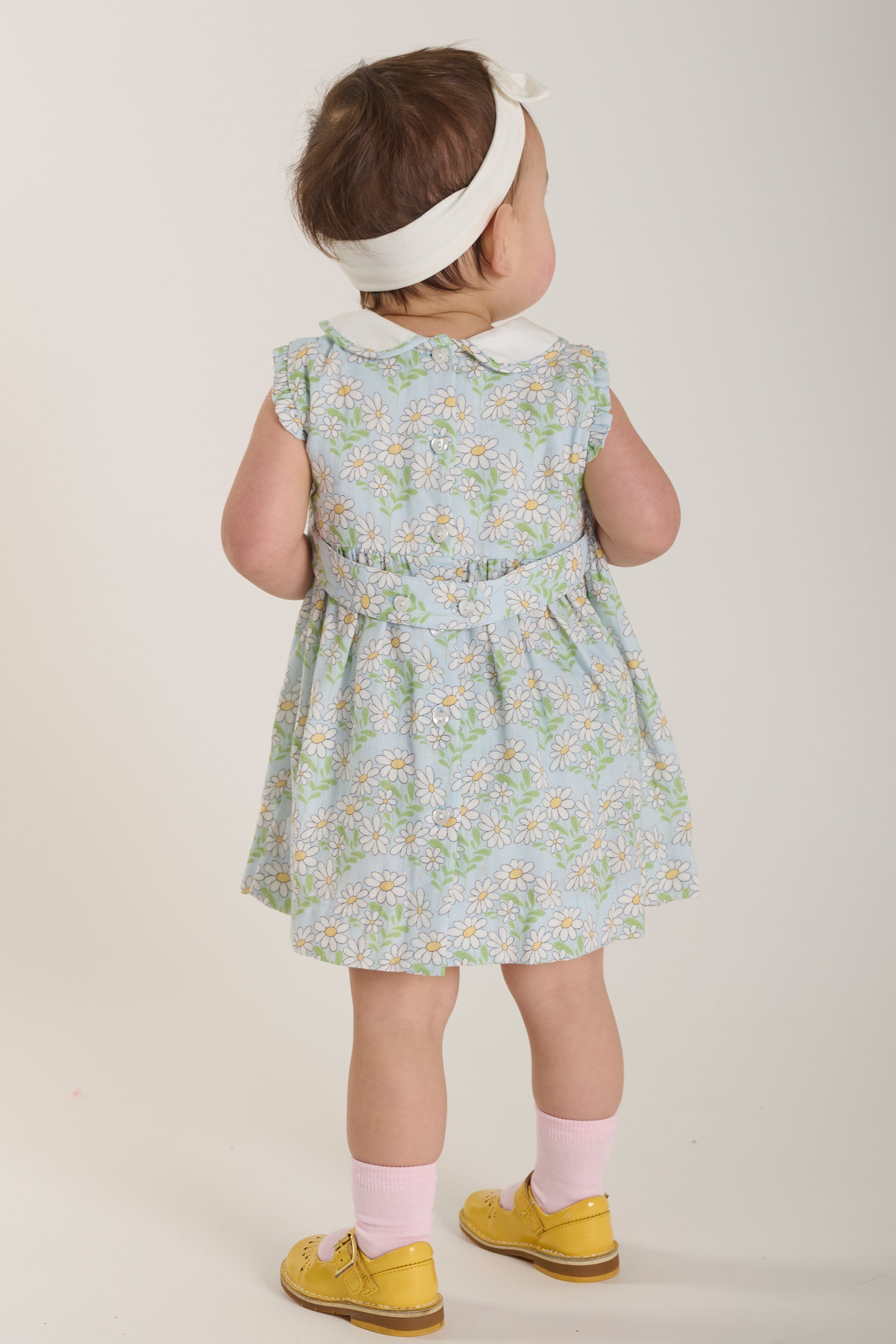 Child wearing a floral dress with a white headband on a plain background seen from behind