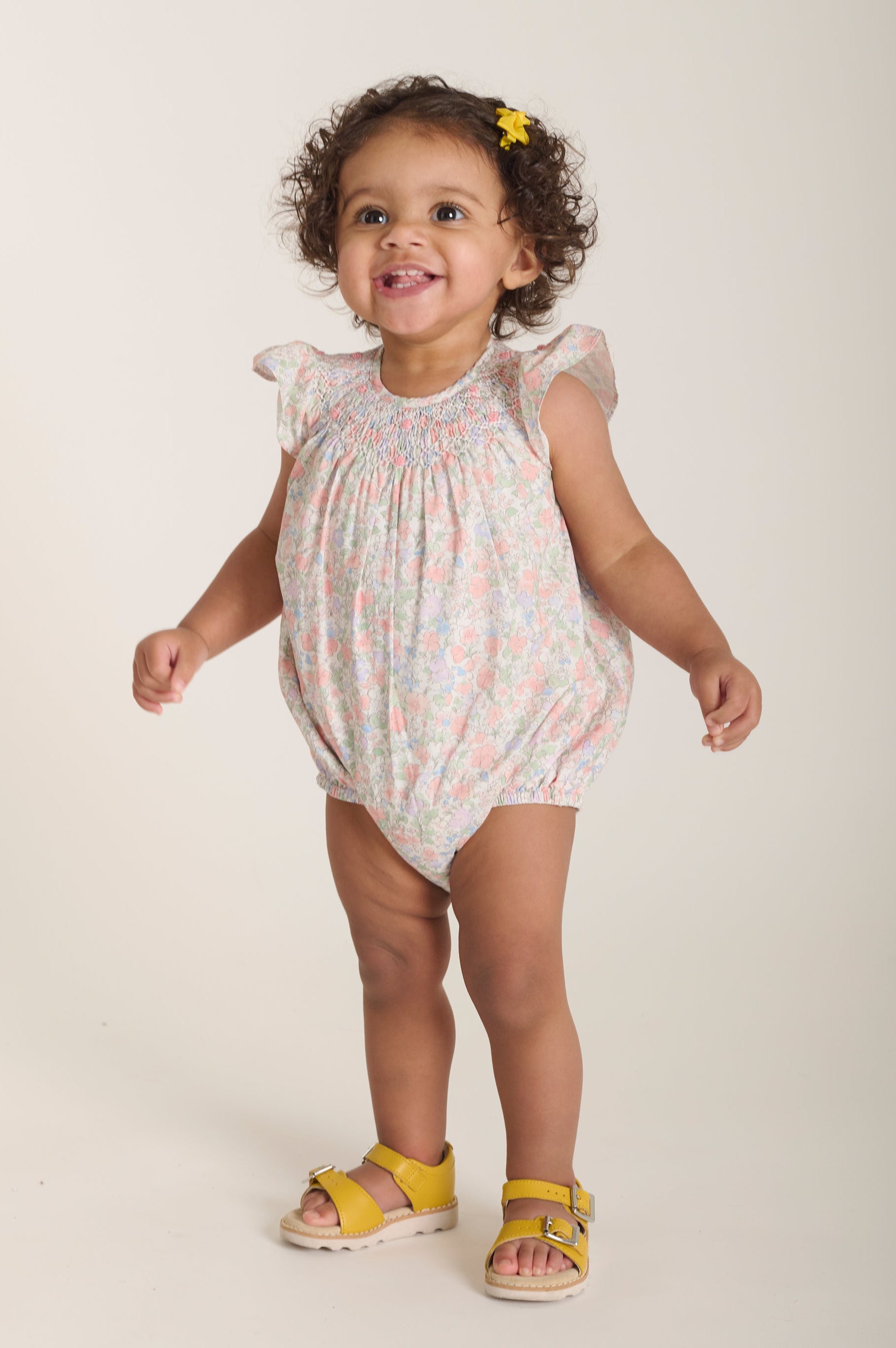 baby wearing a floral romper with yellow sandals on a plain background