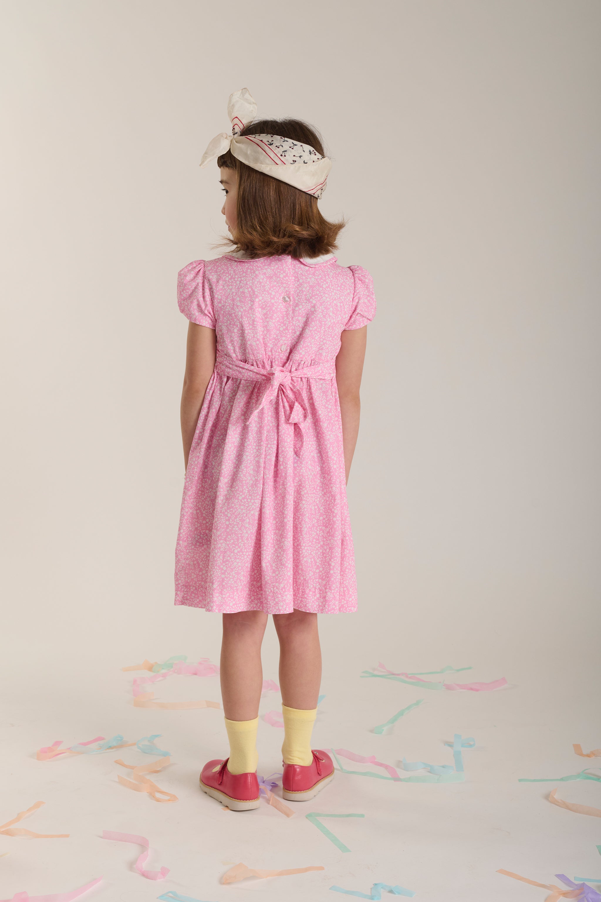 Young girl wearing a pink dress with a belt, standing on a light background with colorful confetti.