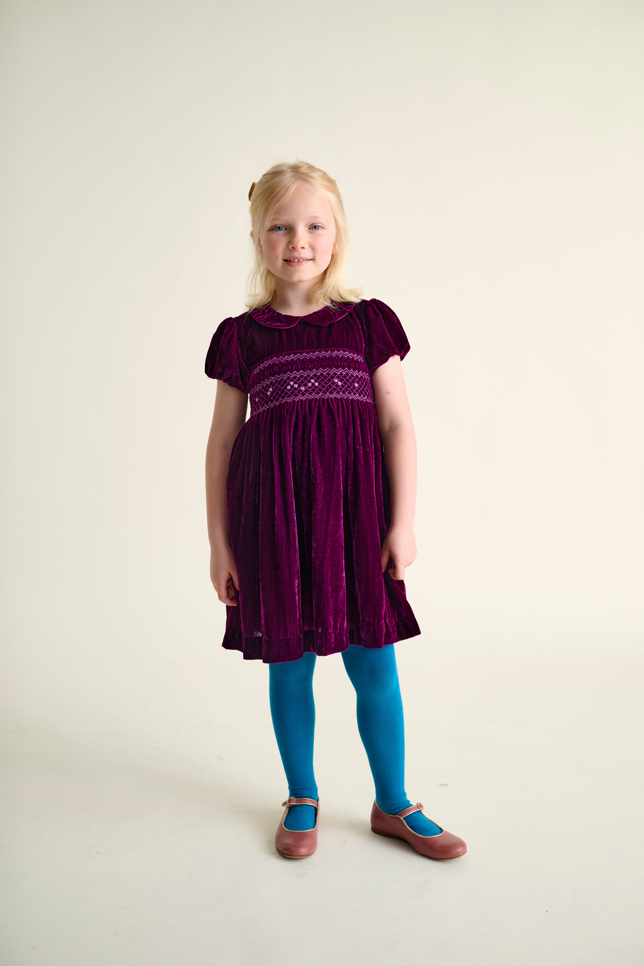 Young girl wearing a burgundy hand-smocked velvet party dress  on a plain background