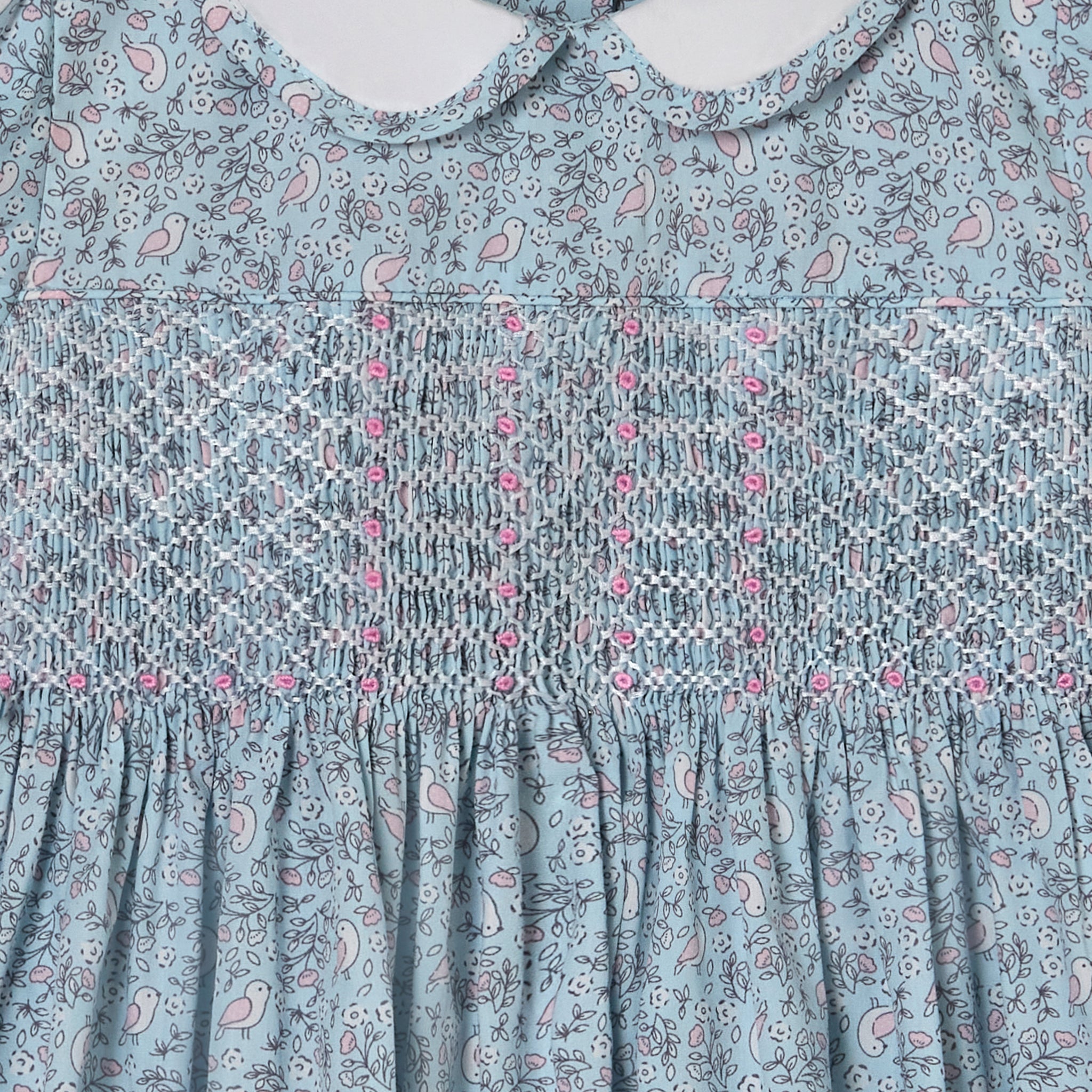 Close-up of a patterned dress with floral and bird design on a light blue background  featuring hand-smocking