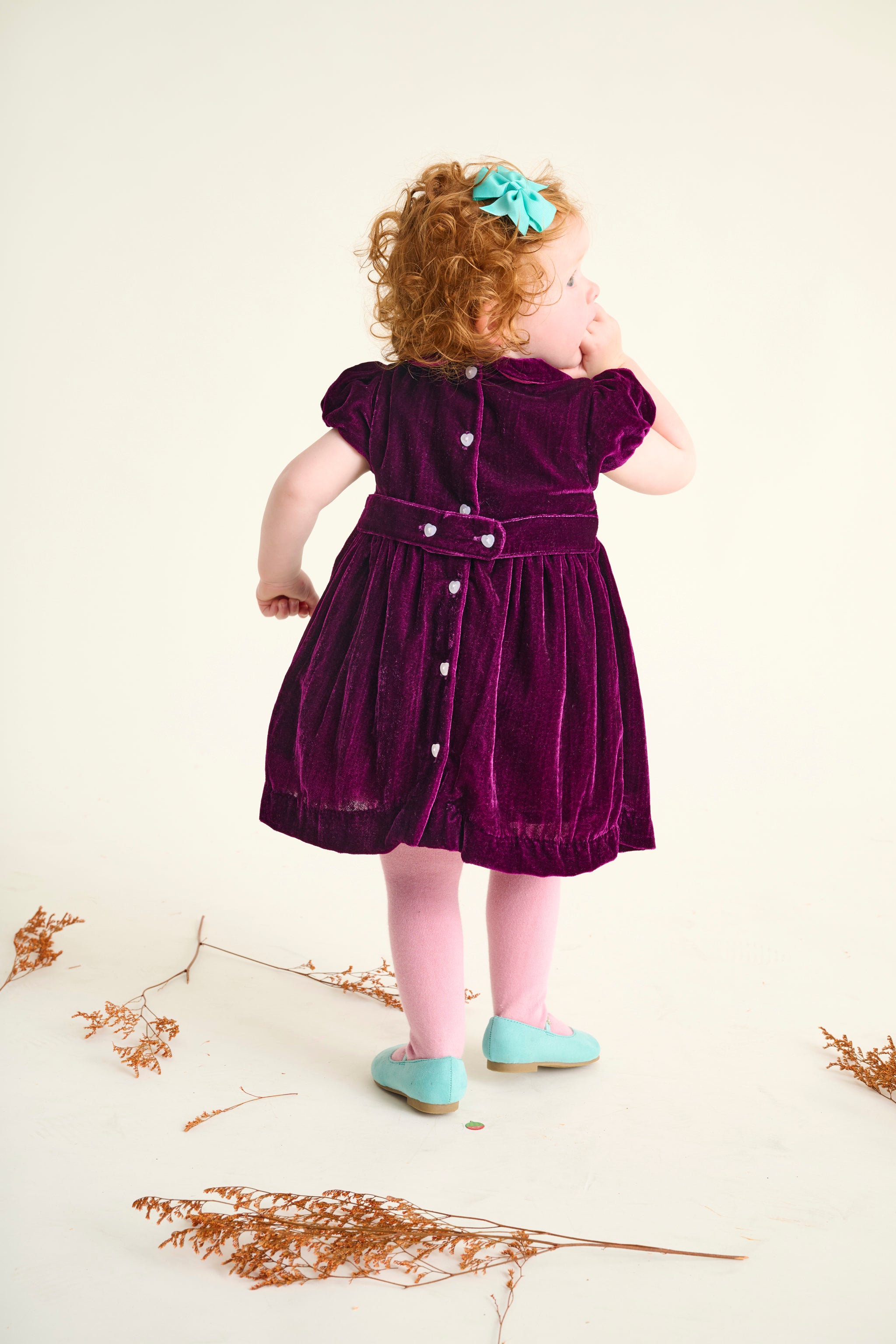 Young girl in a purple velvet dress standing on a light background with dried plants.