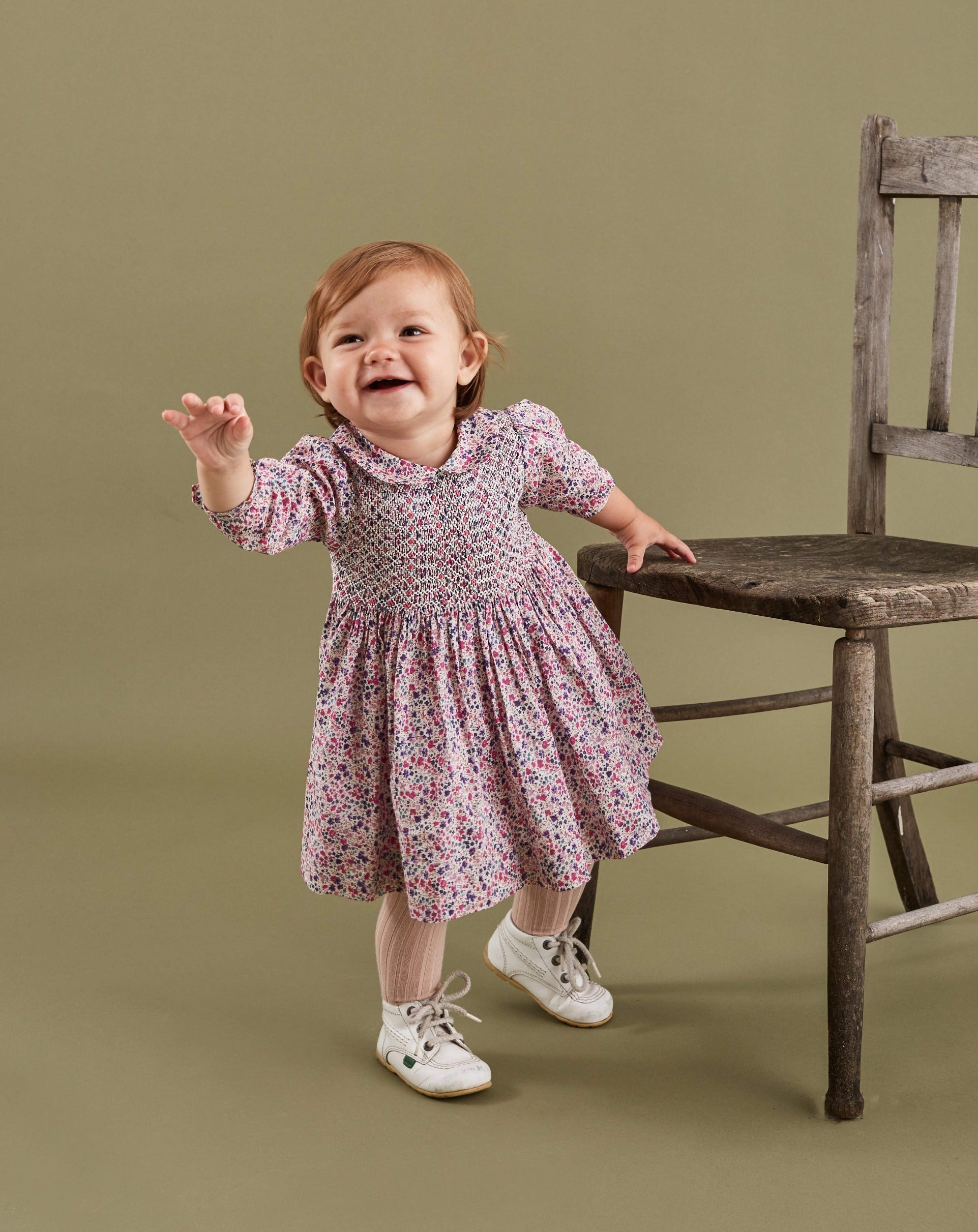 toddler standing next to chair, wearing a floral smocked dress
